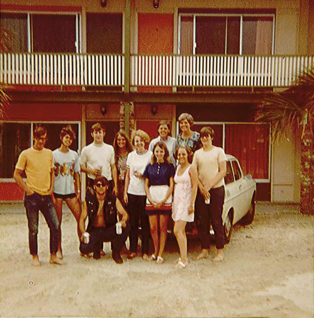 A group of twelve young adults pose in front of a two-story motel and a vintage car, standing on sandy ground on a sunny day. Everyone is casually dressed and smiling.