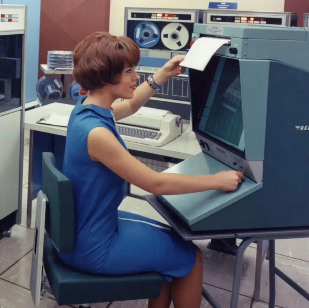 A woman in a blue dress sits at a vintage computer terminal, loading a sheet of paper. Large reel-to-reel tape drives and other computer equipment are visible in the background, indicating a 1960s or 1970s office setting.