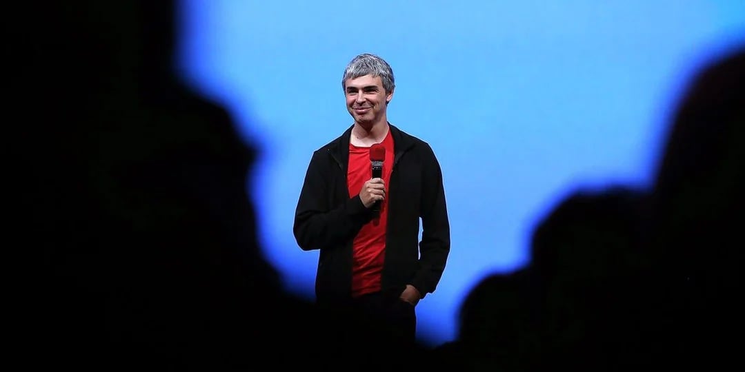 A man with short gray hair, wearing a red shirt and black jacket, stands on stage holding a microphone, smiling, with a blue background and silhouettes of people in the foreground.