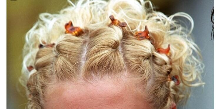 A smiling woman with curly blonde hair styled in small braids decorated with orange hair clips, wearing a grey zip-up sweater, looks directly at the camera.