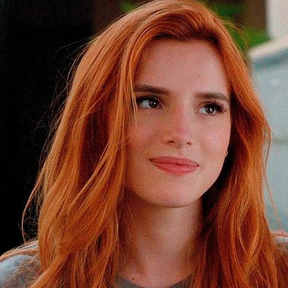 A person with long, wavy red hair and fair skin is smiling softly while looking slightly to the side. They are indoors and wearing a casual gray top. The background is blurred.