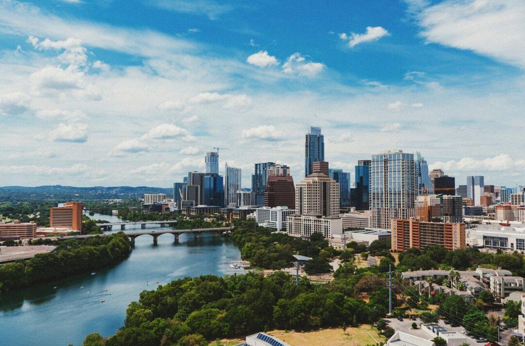 A vibrant city skyline with modern high-rise buildings, a river running through the foreground, a large bridge, and lush green trees under a blue sky with scattered clouds.