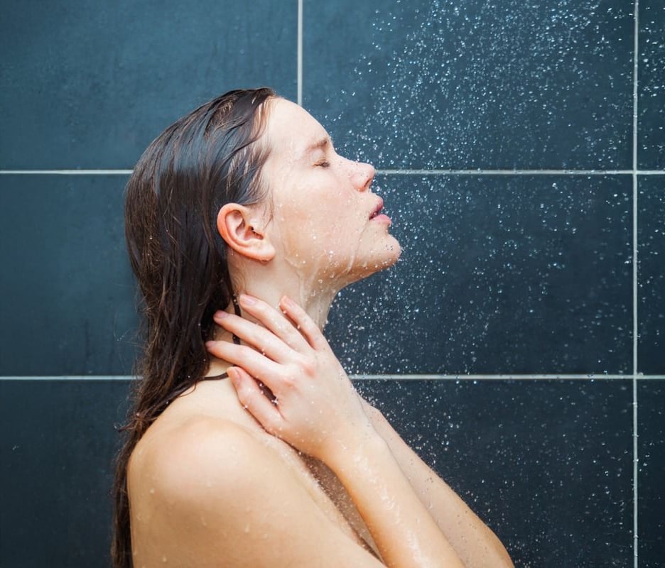 A woman with wet hair stands under a shower with her eyes closed, holding her neck with both hands as water sprays over her face. The background consists of dark tiled shower walls.