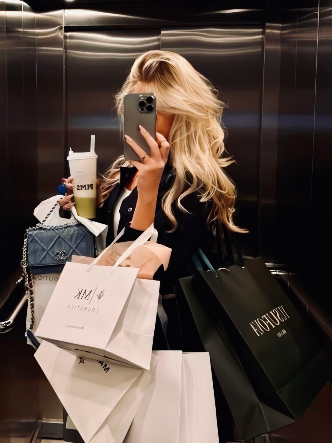 A woman with long blonde hair takes a mirror selfie in an elevator, holding multiple shopping bags, a drink, her phone, and a denim bag, with her face partially covered by her phone.