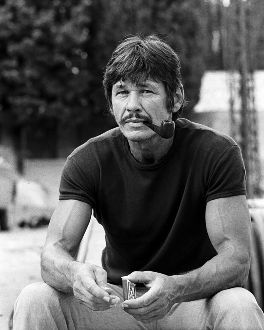 A man with a mustache sits outdoors, wearing a dark t-shirt and holding a pipe in his mouth. He is holding a small rectangular object in one hand and appears relaxed with trees and blurred objects in the background.