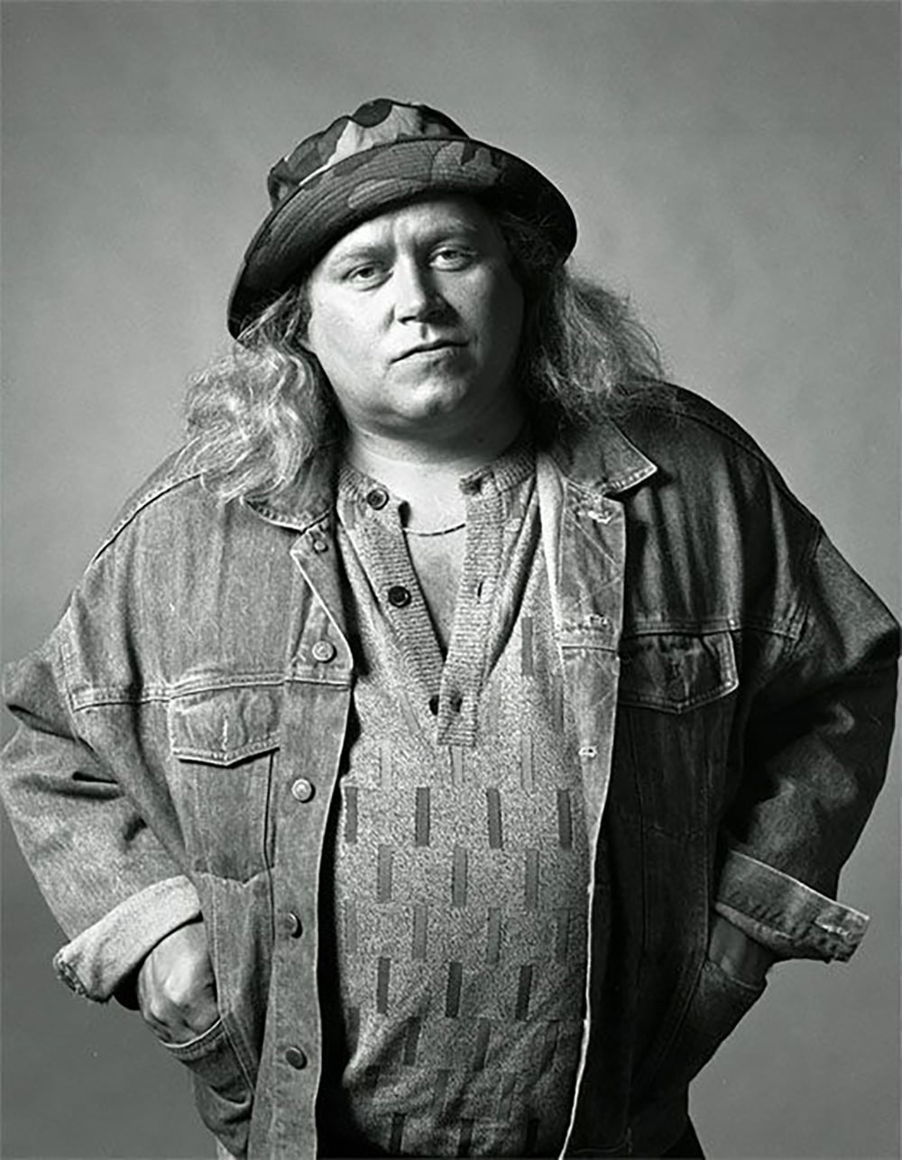 A person with long hair wearing a patterned shirt, a denim jacket, and a bucket hat stands with hands in pockets, looking directly at the camera in a black-and-white portrait.