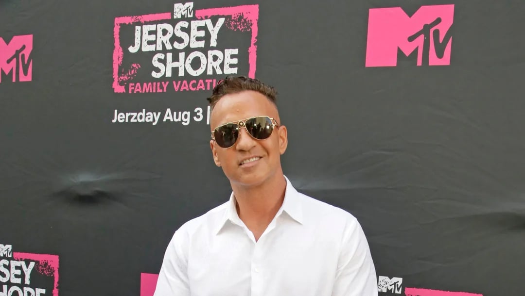 A man in a white shirt and sunglasses smiles in front of a black backdrop with pink MTV and "Jersey Shore Family Vacation" logos and text promoting the show.