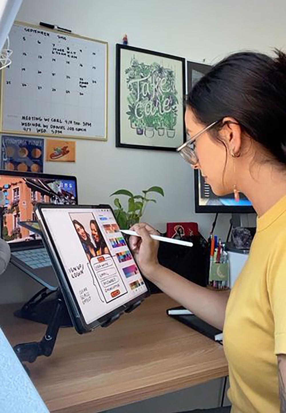 A woman wearing glasses and a yellow shirt draws on a tablet with a stylus at a desk, with a computer monitor, plant, wall calendar, and framed art visible in the background.