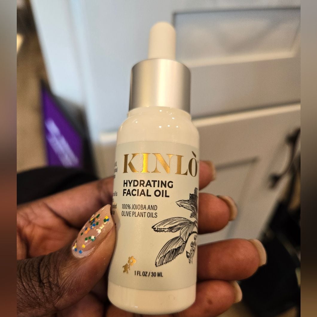A hand with glittery nail polish holds a bottle of KINLÒ Hydrating Facial Oil, labeled as containing 100% jojoba and olive plant oils. The bottle is white with gold and black lettering and a dropper cap.