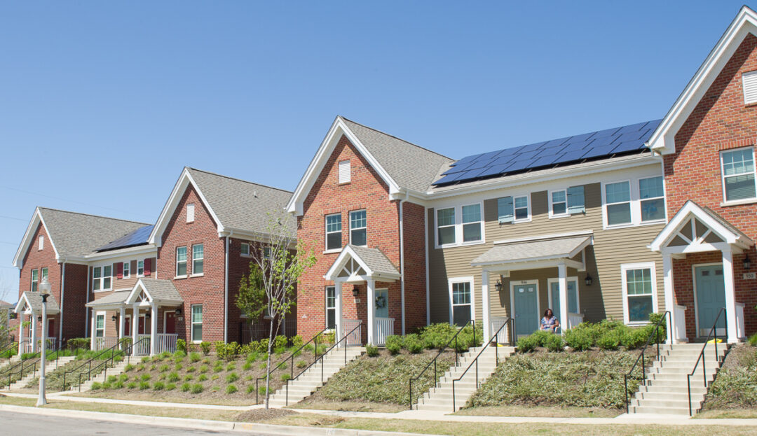 A row of modern townhouses with brick and siding exteriors. One house has solar panels on the roof. A person sits on the front steps. The sky is clear and blue, and young trees and shrubs line the sidewalk.