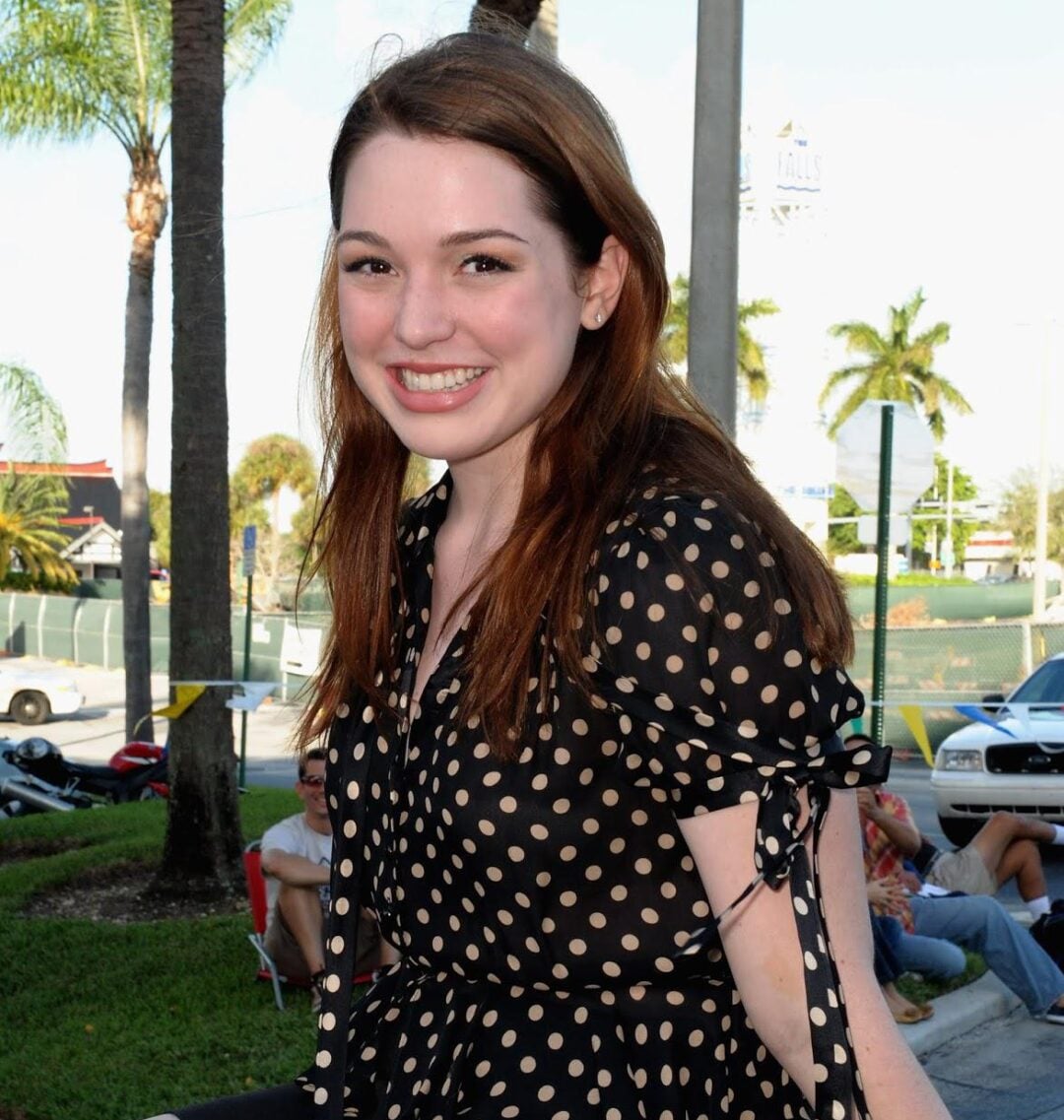 A young woman with fair skin and long brown hair sits outdoors, smiling at the camera. She wears a black blouse with white polka dots and black shorts. Palm trees and parked cars are visible in the background.