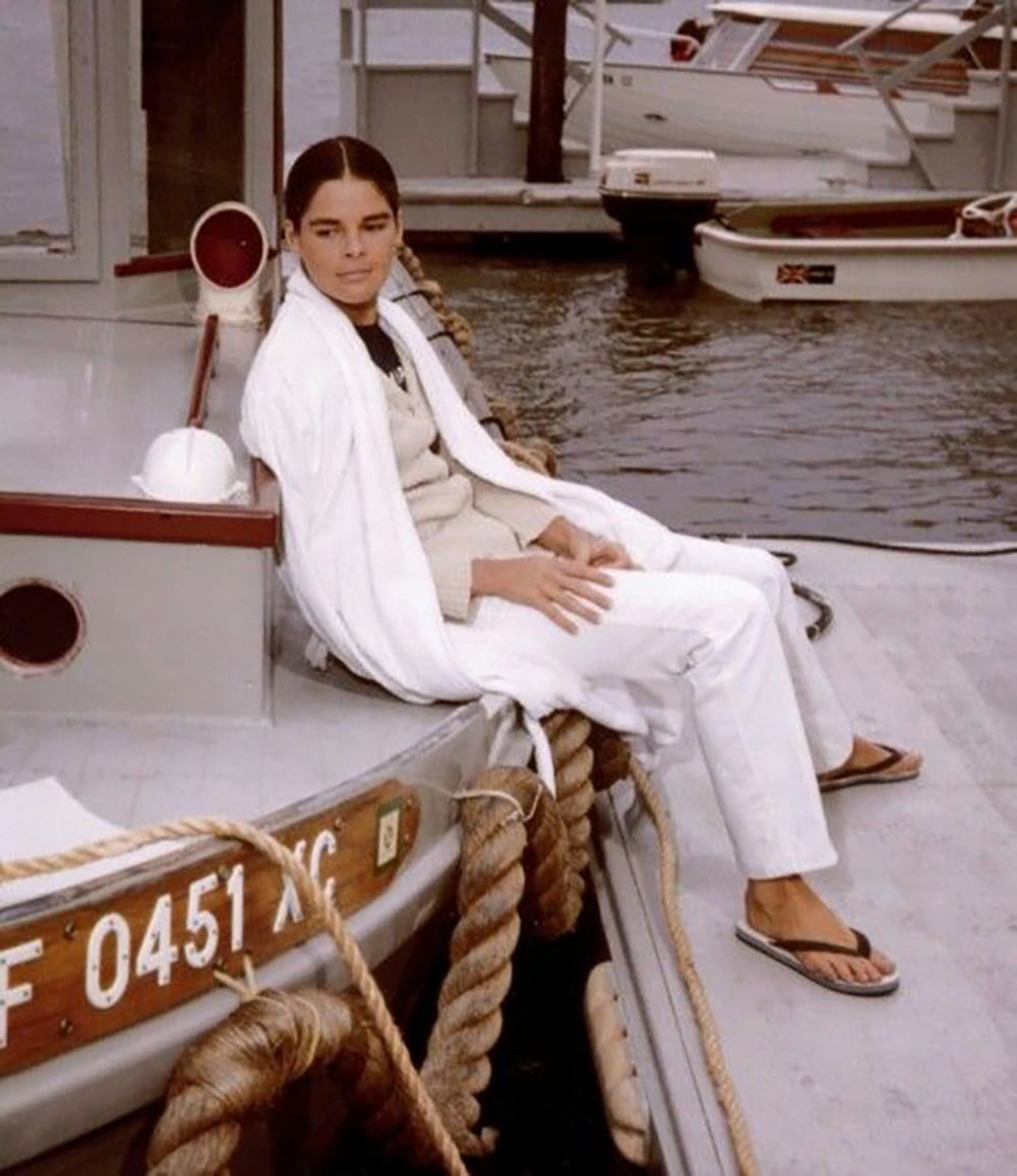 A person wearing white clothes and a bathrobe sits on a docked boat, with more boats in the background. They have dark hair pulled back and are wearing flip-flops, looking relaxed by the water.