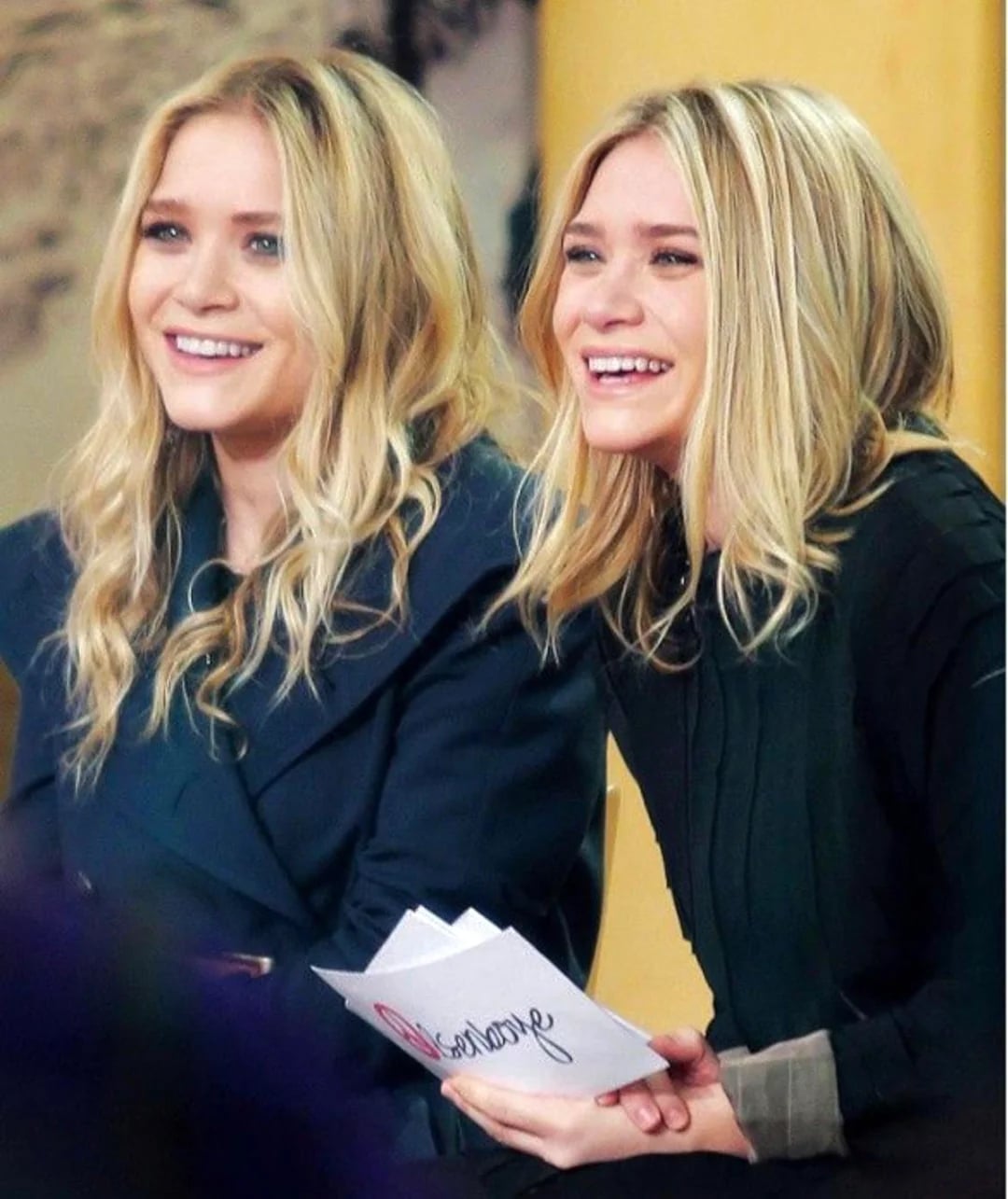 Two young women with long blonde hair sit side by side, smiling and looking slightly to the left. One is holding a booklet or papers. Both are wearing dark-colored outfits in a brightly lit indoor setting.