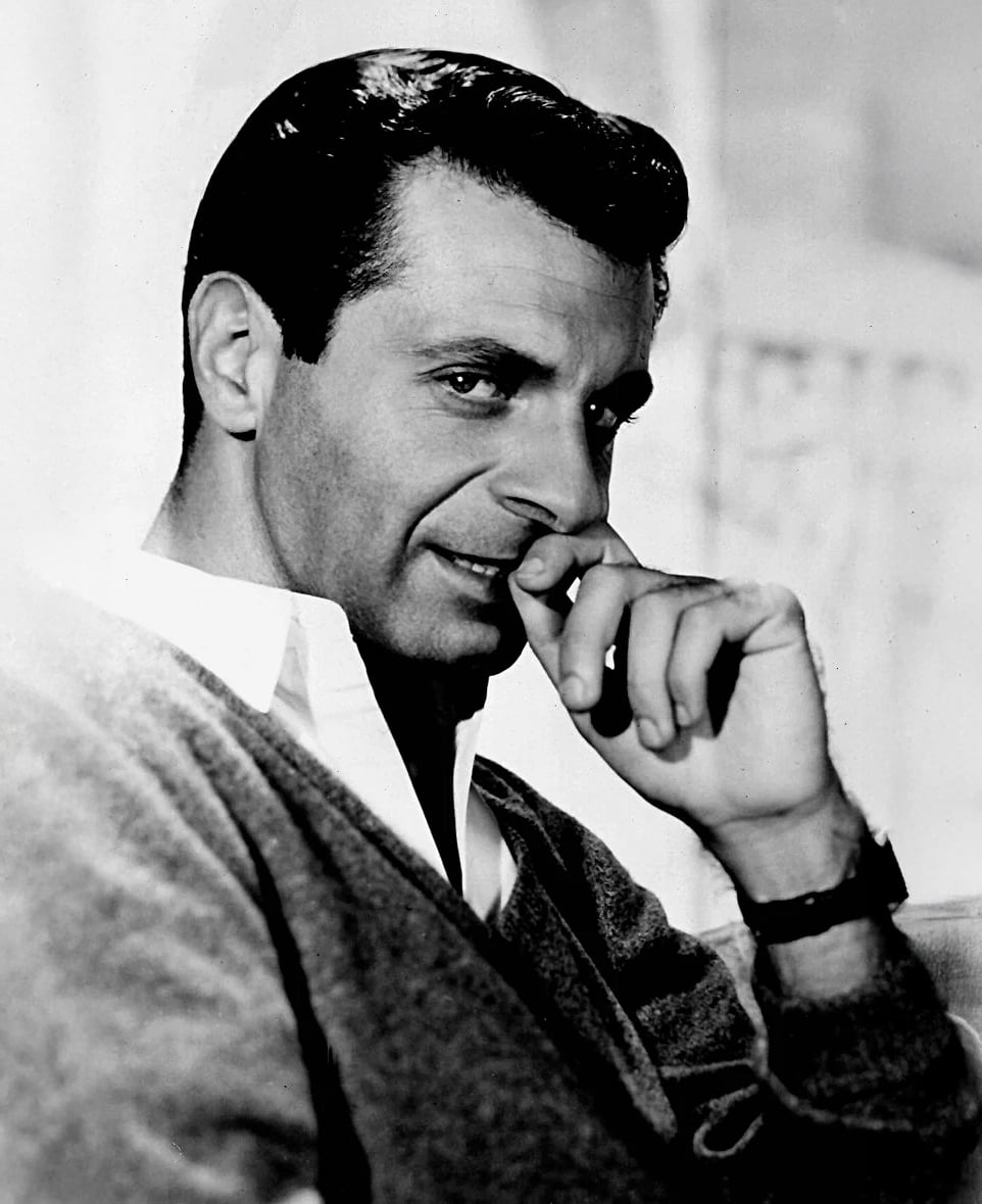 Black-and-white portrait of a man with dark hair, wearing a collared shirt and sweater, sitting and looking at the camera with a slight smile while resting his hand on his chin.
