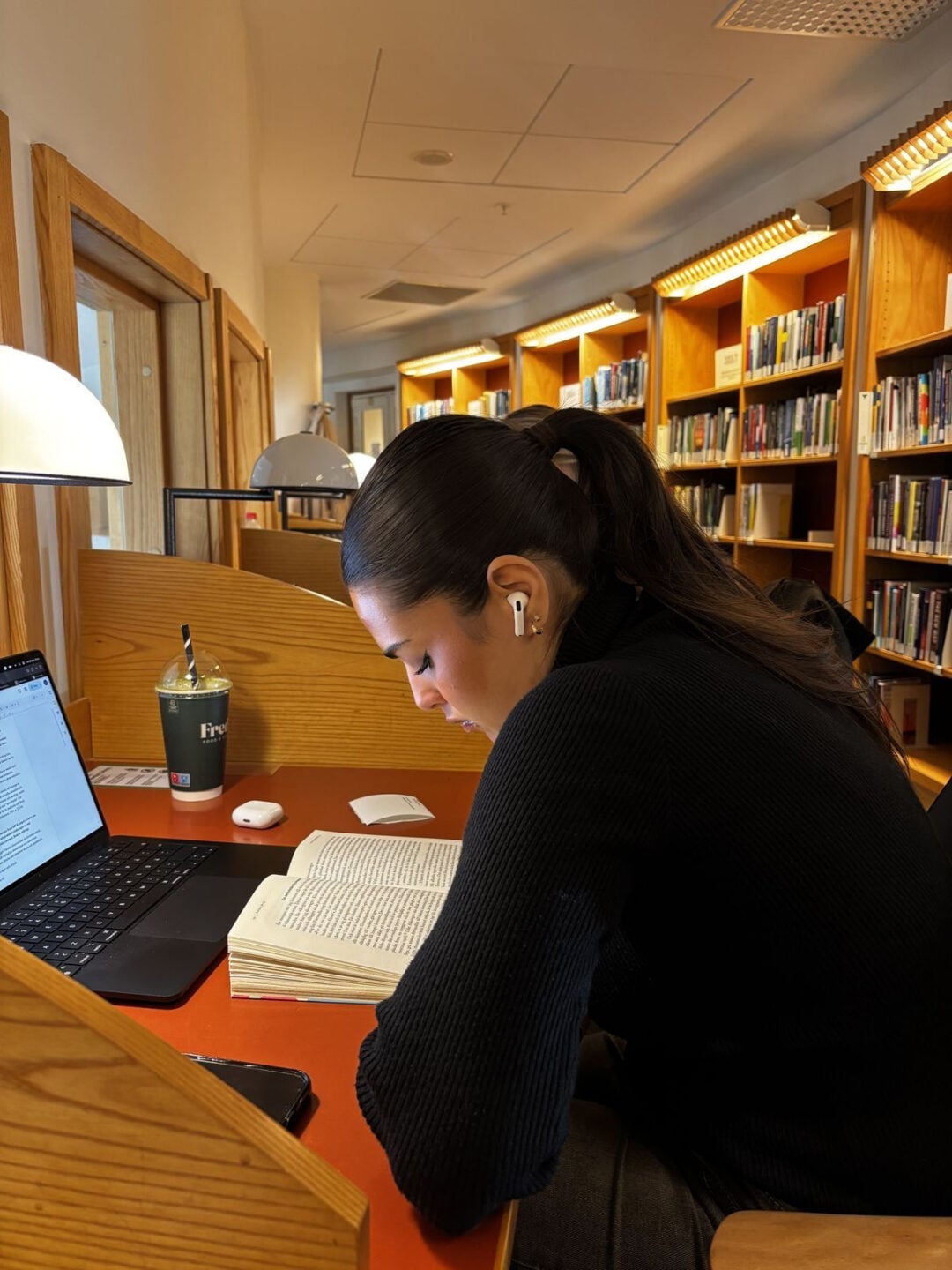 A person with dark hair in a ponytail reads a book at a library desk with a laptop, phone, drink, and mouse nearby. Bookshelves and desk lamps are in the background.