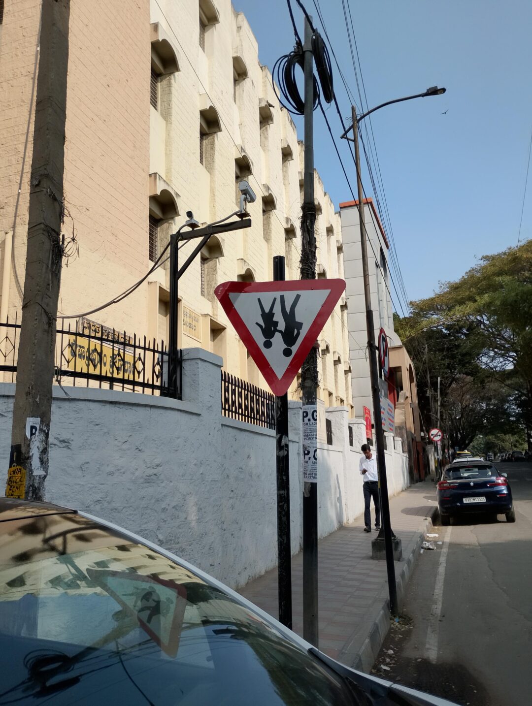 A triangular street sign with an image of two cows facing each other stands on a sidewalk beside a building; cars are parked along the road and a person is walking in the background.