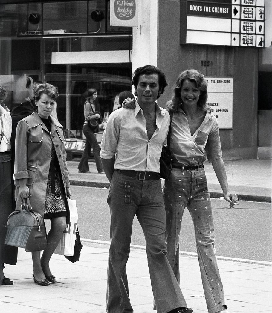 A man and woman walk arm in arm on a city sidewalk, both smiling. The man wears a partially unbuttoned shirt and flared trousers; the woman wears a blouse and polka-dot flared pants. Other pedestrians walk nearby. The photo is in black and white.