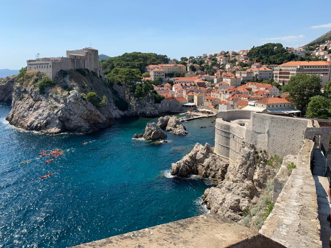 View of Dubrovnik’s old town in Croatia, with historic stone walls, orange rooftops, and a fortress on a rocky cliff by the turquoise Adriatic Sea. Kayakers paddle near the shore under a clear blue sky.