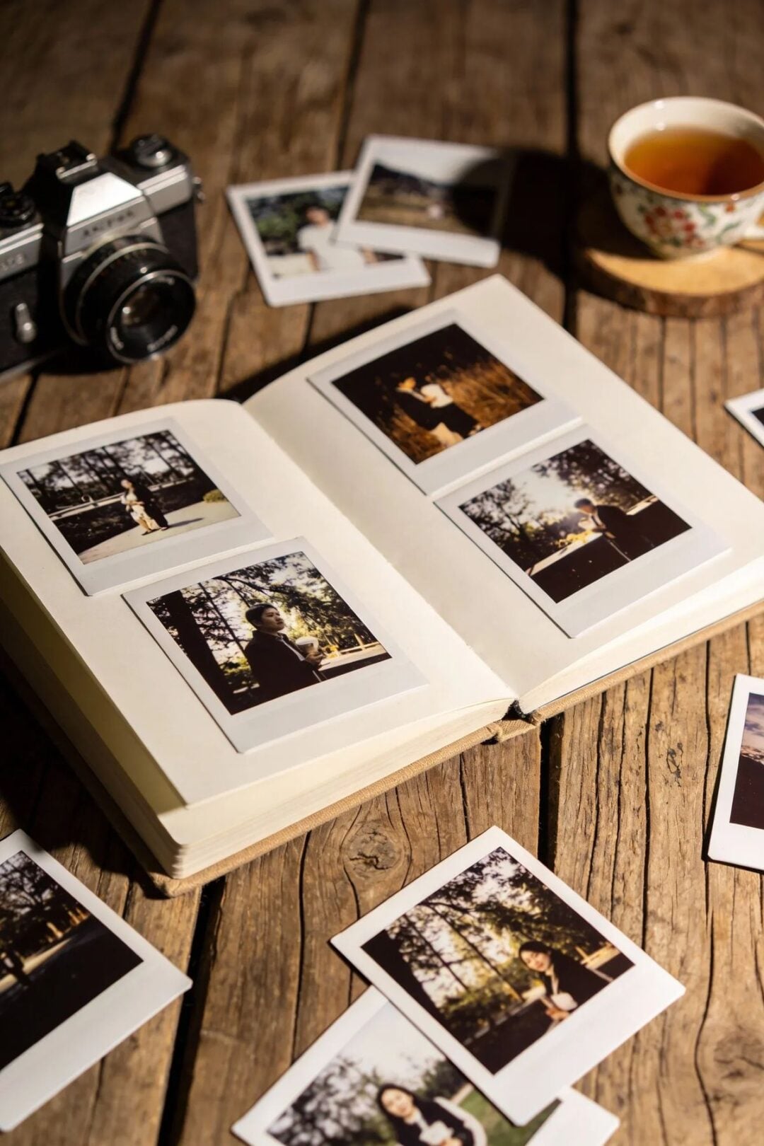 An open photo album with instant photos on a wooden table, surrounded by more loose photos, a vintage camera, and a teacup filled with tea. Warm lighting suggests a cozy, nostalgic atmosphere.