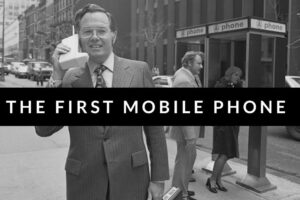 A man in a suit holds a large, early mobile phone to his ear on a city street, with payphones and pedestrians visible in the background. The text "THE FIRST MOBILE PHONE" is overlaid on the image.