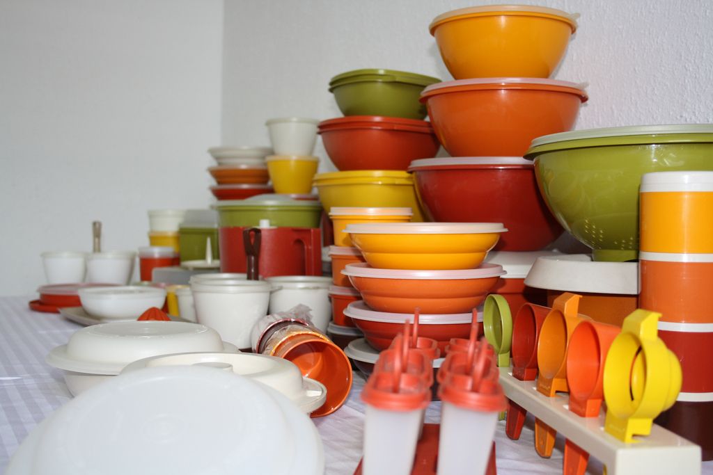 A collection of colorful vintage plastic kitchenware, including bowls, cups, plates, and containers, arranged neatly on a table against a white wall. Most items are orange, yellow, olive green, and white.