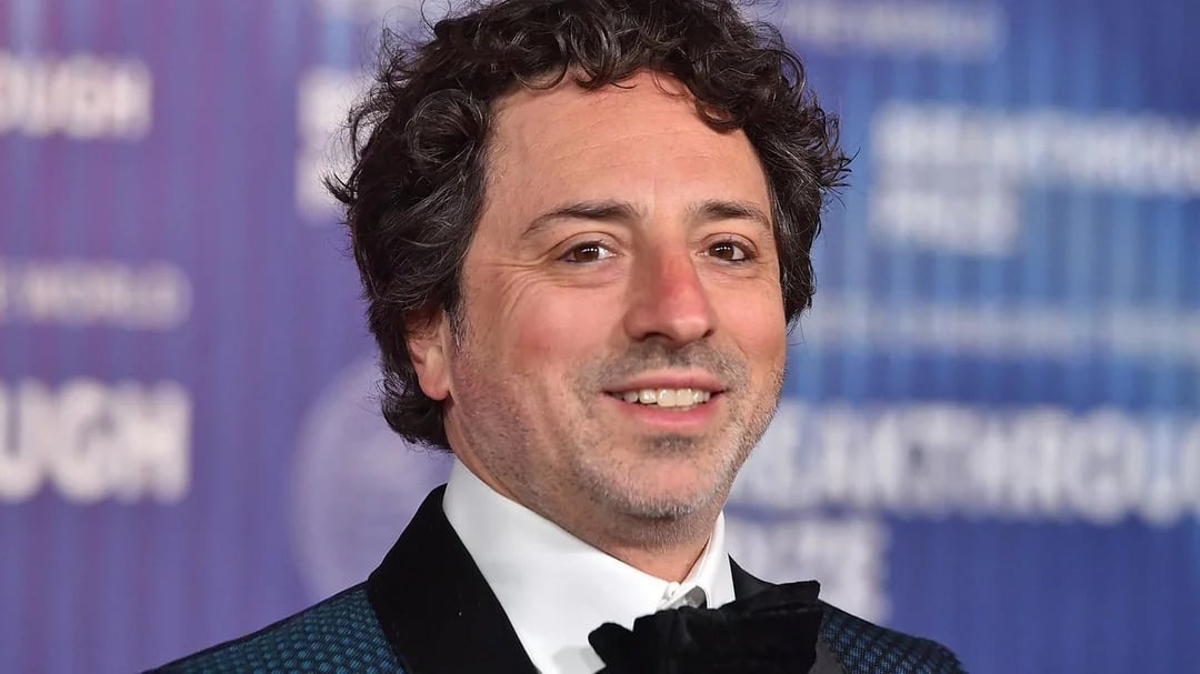 A man with curly dark hair and a short beard wears a dark tuxedo and bow tie, smiling at an event with a blue background featuring blurred white text.