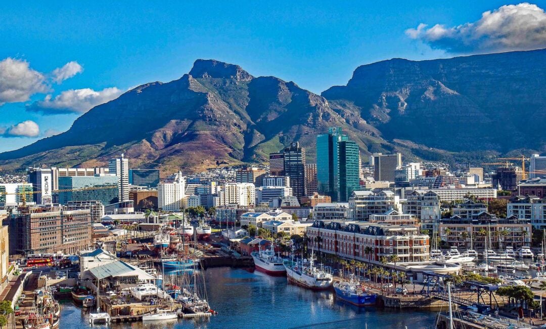 A vibrant cityscape of Cape Town, South Africa, with tall buildings, boats docked in the harbor, and the iconic Table Mountain and Lion’s Head towering in the background under a clear blue sky.