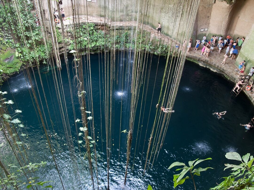 A large, circular cenote with deep blue water is surrounded by stone and lush vegetation. Long vines hang down into the cenote, while people swim and stand along the edge, some preparing to jump in.