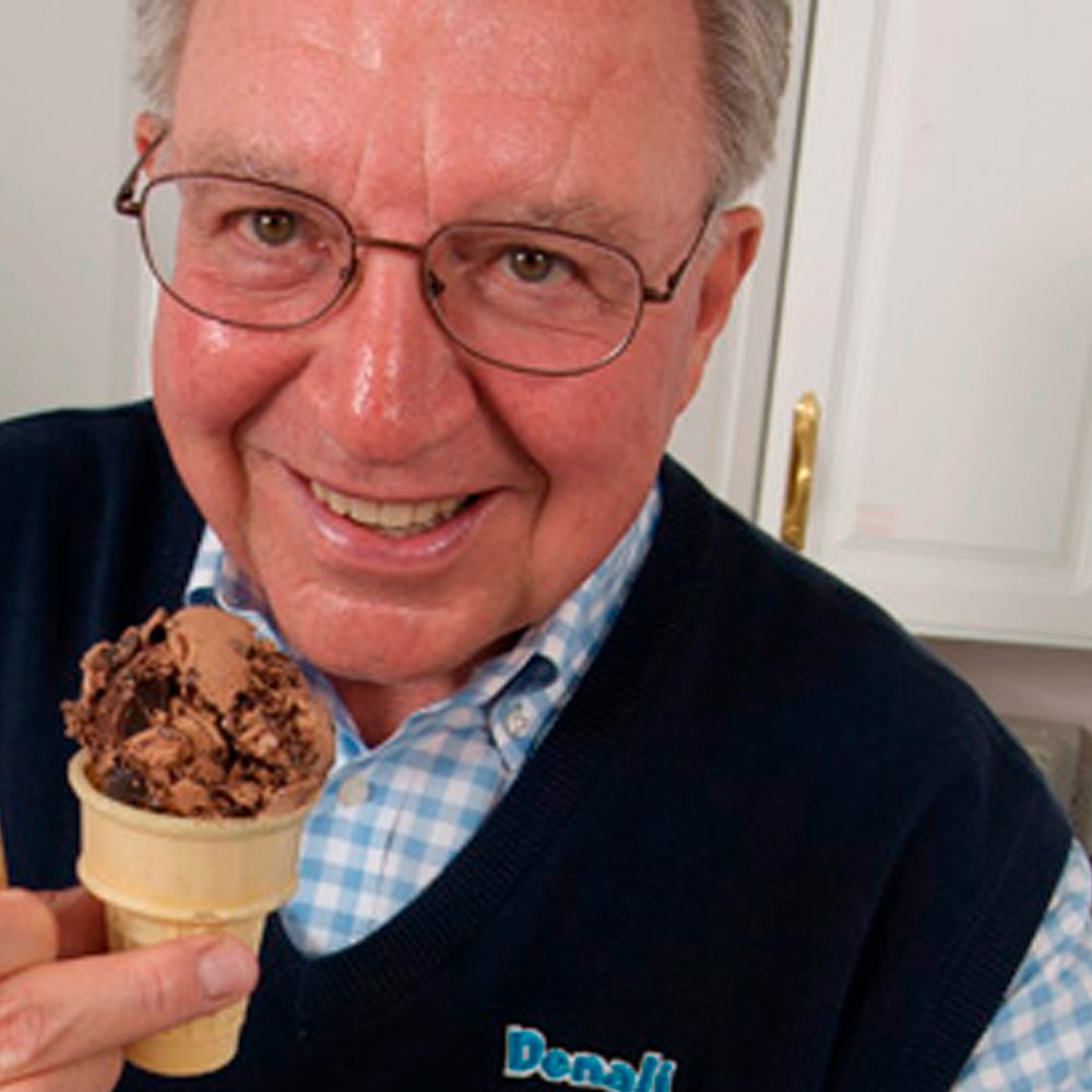 An older man wearing glasses and a blue sweater smiles while holding a chocolate ice cream cone in a kitchen.