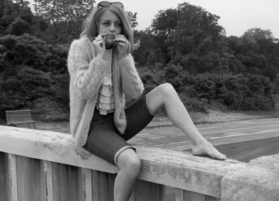A young woman with long hair sits barefoot on a concrete fence, wearing a cardigan, ruffled blouse, and shorts, and eats a sandwich. Trees and a roadway are visible in the background.