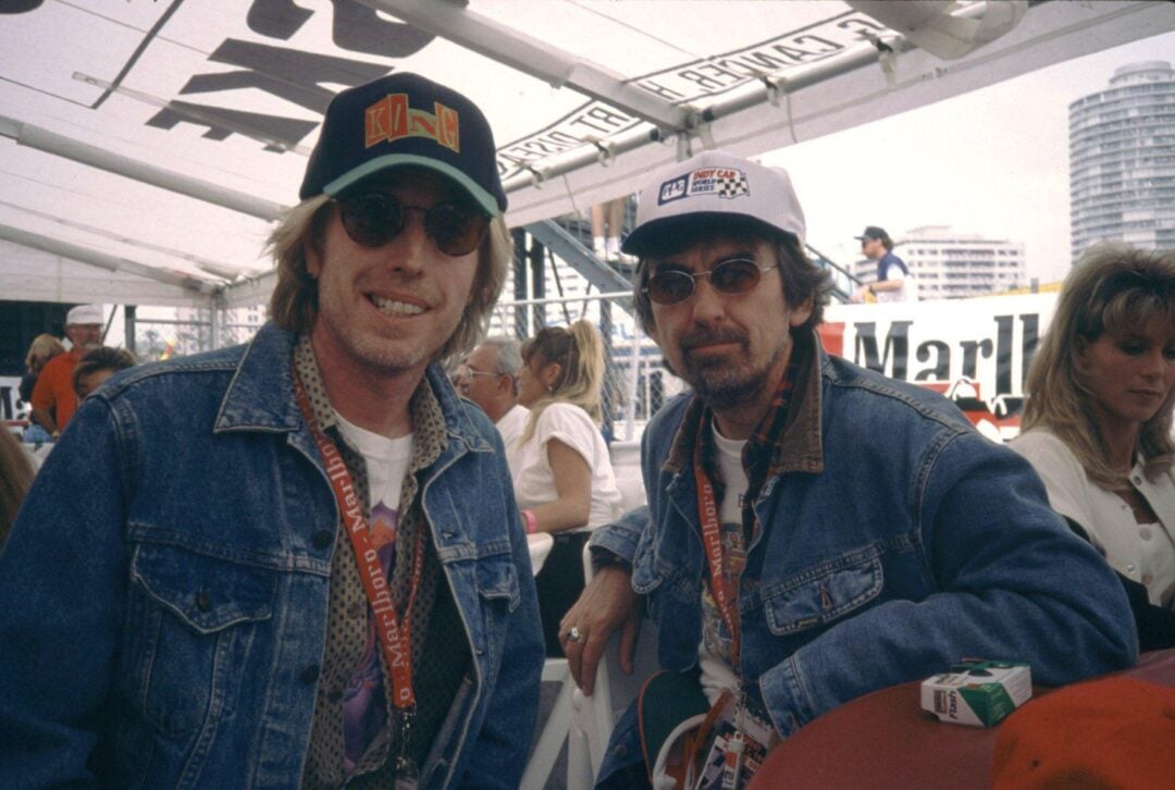 Two men wearing denim jackets, sunglasses, and caps pose and smile at a crowded outdoor event. They both have event lanyards around their necks. There are people and city buildings in the background.