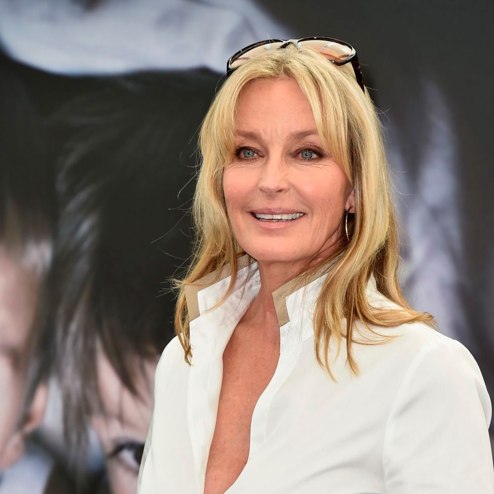 A smiling woman with long blonde hair, wearing a white collared shirt and sunglasses on her head, stands in front of a blurred dark background.