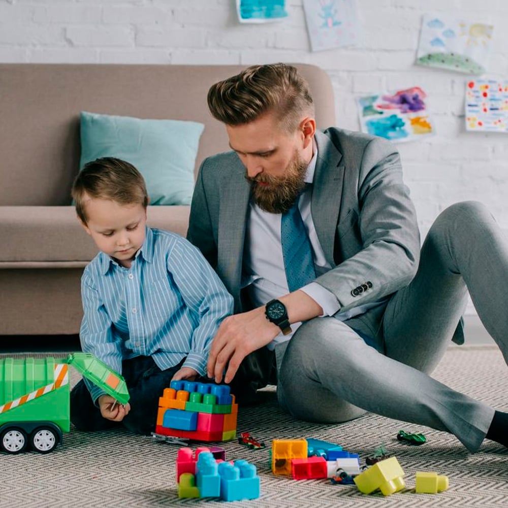 A man in a suit sits on the floor beside a young boy, helping him build with colorful plastic blocks. They are in a living room with a sofa, cushions, and children's drawings on the white brick wall.