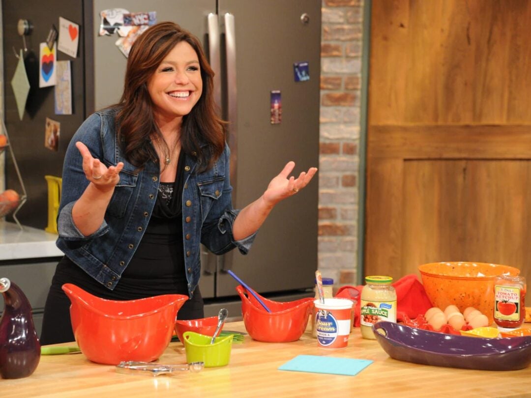 A woman smiles and gestures with her hands in a kitchen, standing behind a counter with colorful mixing bowls, ingredients, and utensils. A fridge and wooden cabinets are visible in the background.