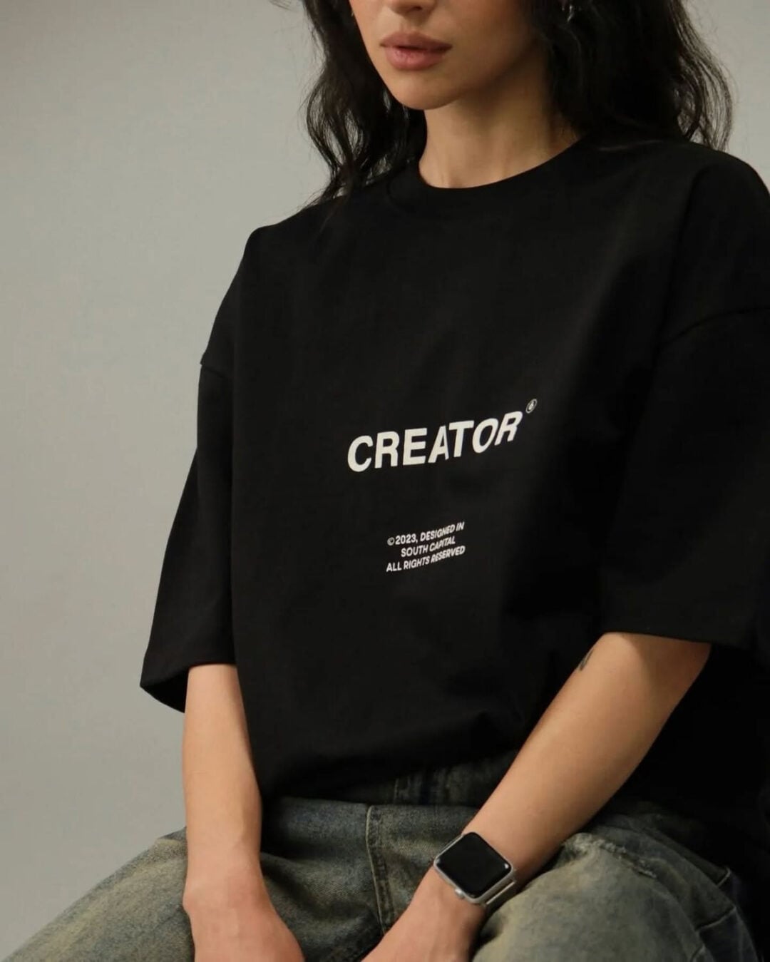 A woman with long dark hair is wearing a black oversized T-shirt that says "CREATOR" in white text, along with additional small writing below. She is also wearing a smartwatch and sitting with her hands in her lap.