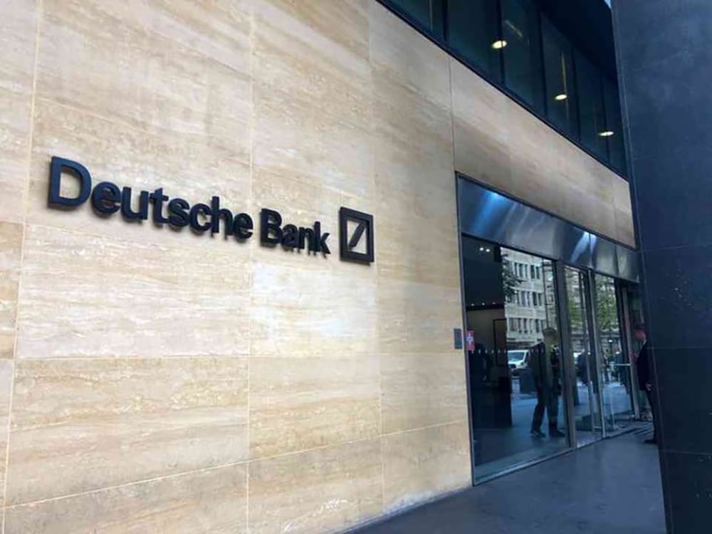 The exterior of a Deutsche Bank branch, showing the bank’s name and logo on a beige stone wall next to glass doors through which people can be seen inside.