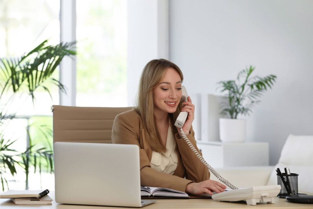 A woman in a tan blazer sits at a desk with a laptop, open notebook, and pen holder, smiling while talking on a landline phone in a bright, modern office with green plants in the background.