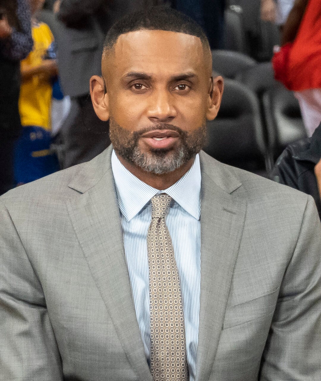 A man in a gray suit and striped tie sits at a desk with an NBA TV sign and a laptop, appearing to be on a sports broadcast set.