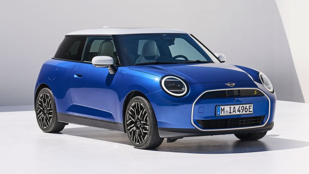 A blue Mini Cooper electric car with a white roof is parked on a smooth white surface, facing left. The car features black wheels, sleek curves, and modern headlights, with a European license plate on the front.