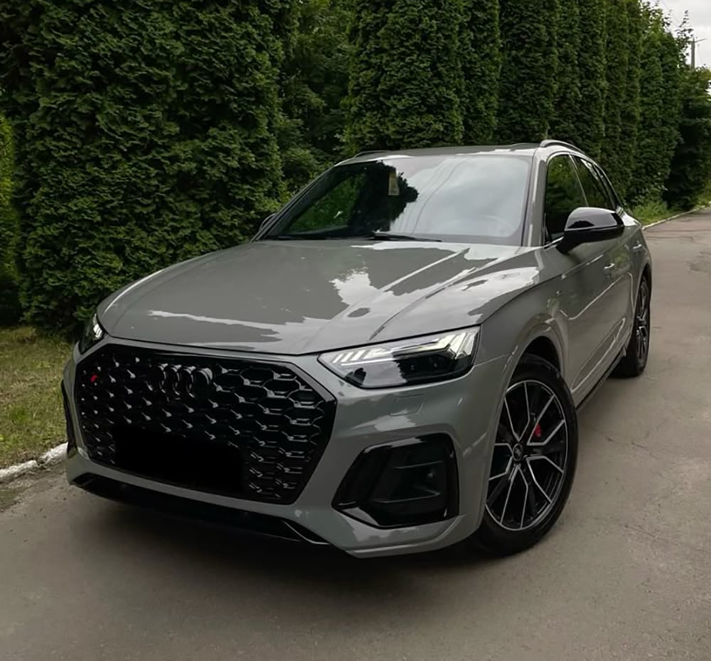 A gray Audi SUV is parked on a paved road next to tall green hedges on a cloudy day.
