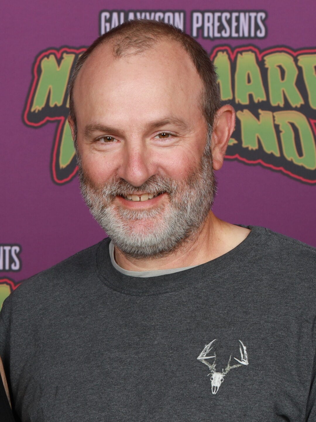 A middle-aged man with a gray beard and short hair smiles at the camera. He is wearing a dark shirt with a white deer skull graphic, standing in front of a purple backdrop with colorful text.