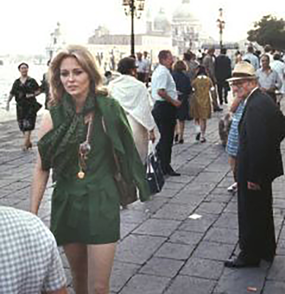 A woman in a green outfit walks on a crowded waterfront promenade, with people strolling and historic buildings visible in the background on a hazy day.