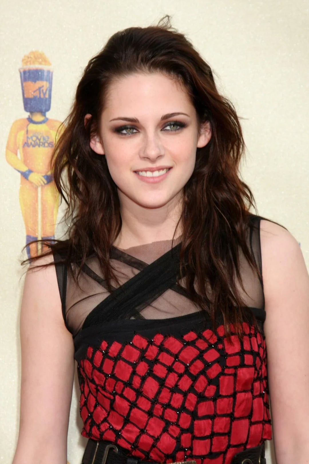 A woman with long, wavy brown hair wearing a red and black patterned dress smiles while standing in front of a backdrop with a gold MTV Movie Awards statue.