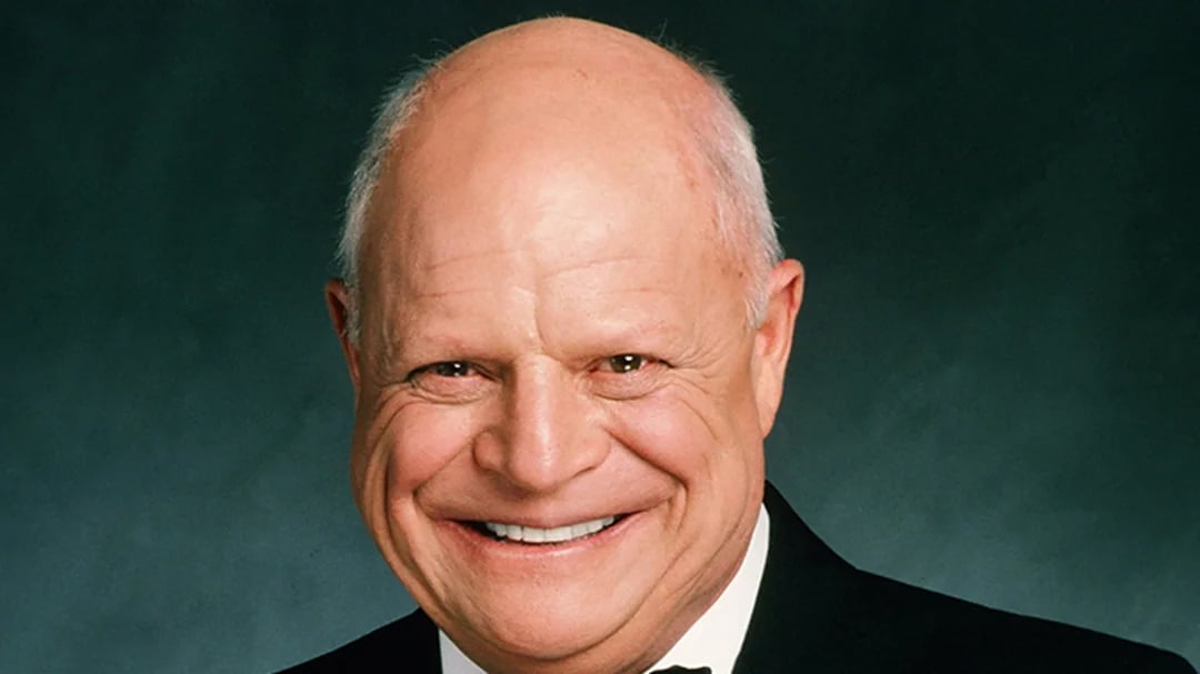 A smiling older man with a bald head wearing a black tuxedo, white shirt, and black bow tie, posed in front of a plain dark background.