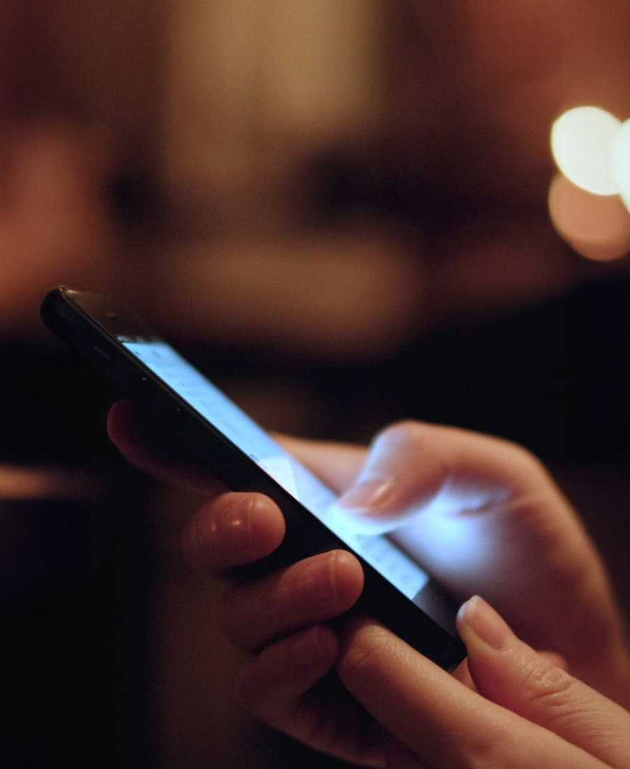 A close-up of a person's hands holding and using a smartphone, with the screen glowing in a dimly lit environment.