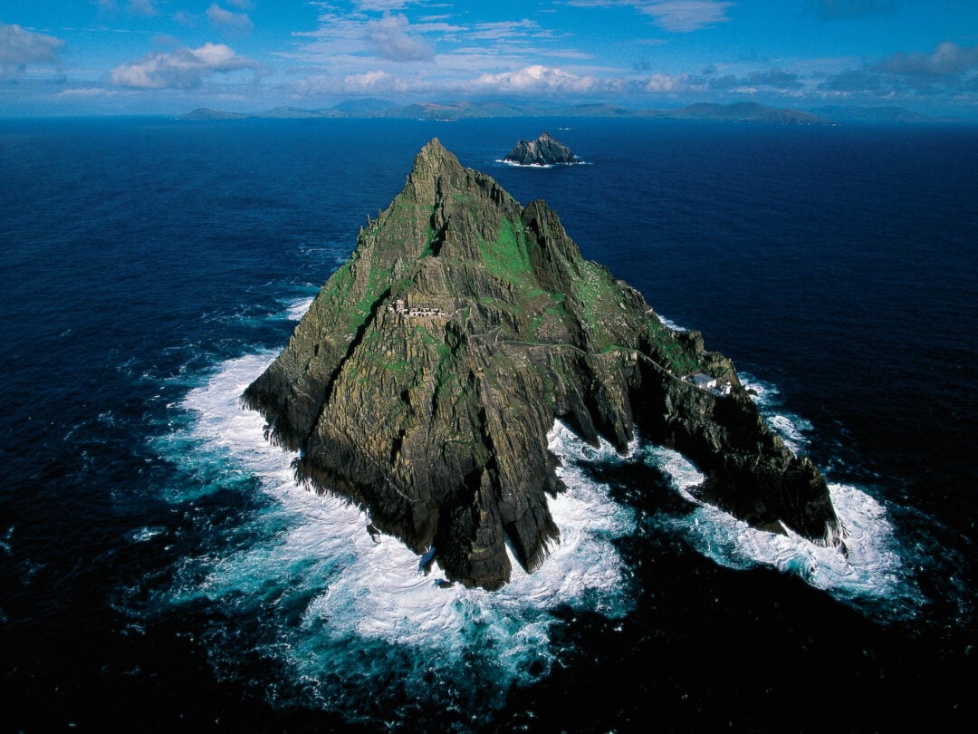 A rocky, steep island covered in patches of green rises from the deep blue ocean, surrounded by white waves crashing against its base. A smaller island is visible in the distance under a partly cloudy sky.
