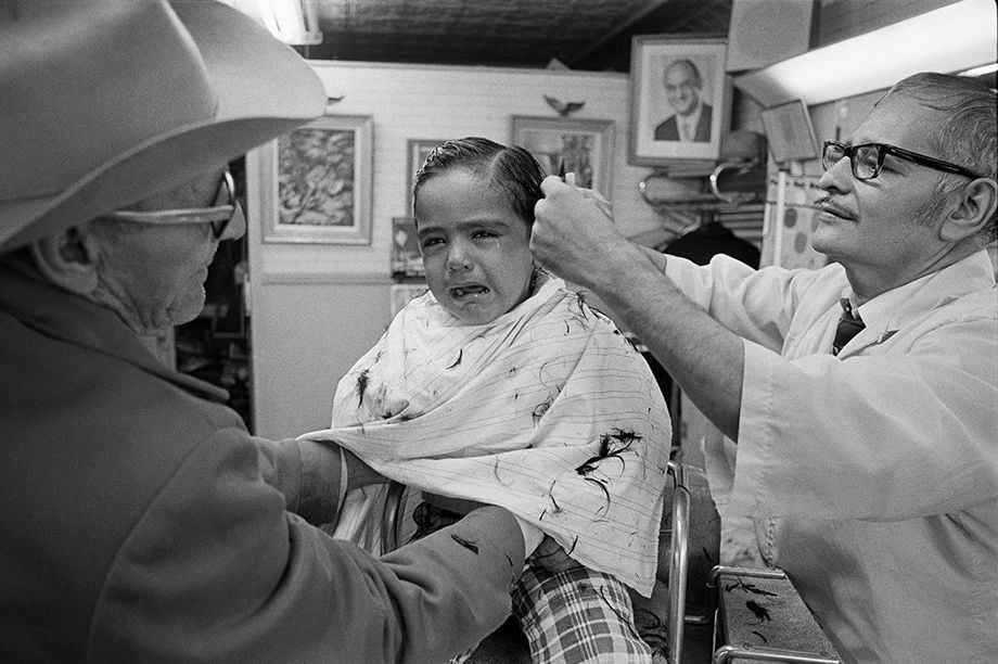 A young child sits in a barber chair, crying while getting a haircut. An older man comforts the child as the barber cuts their hair. Hair clippings are visible on the child's cape. Framed photos hang on the walls in the background.