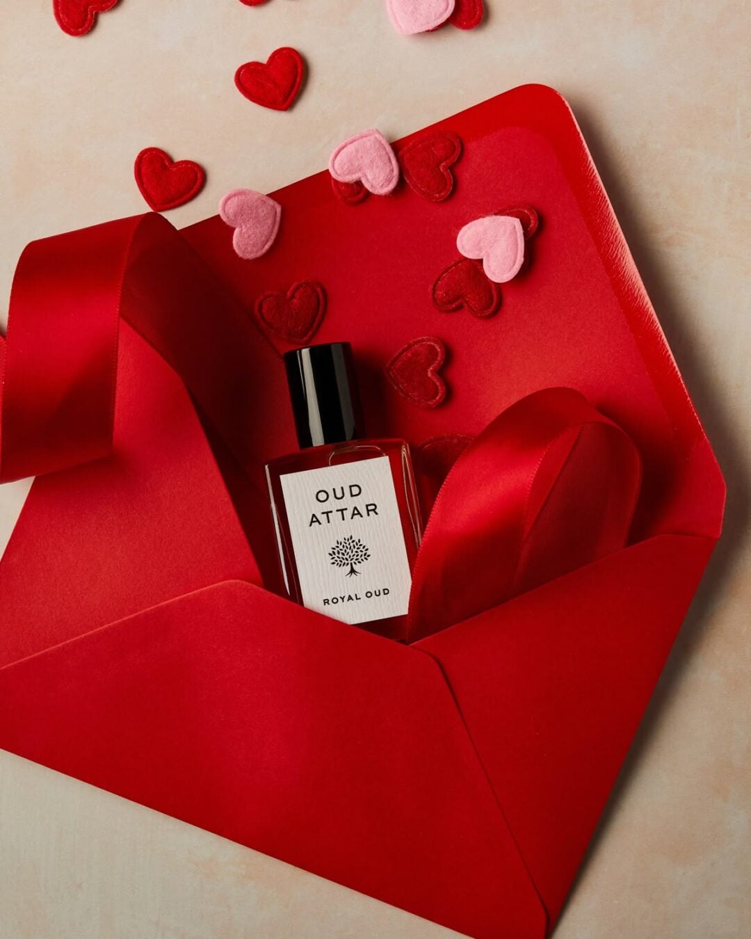 A bottle of Oud Attar Royal Oud perfume in a red envelope with a red ribbon, surrounded by red and pink felt hearts on a light surface.