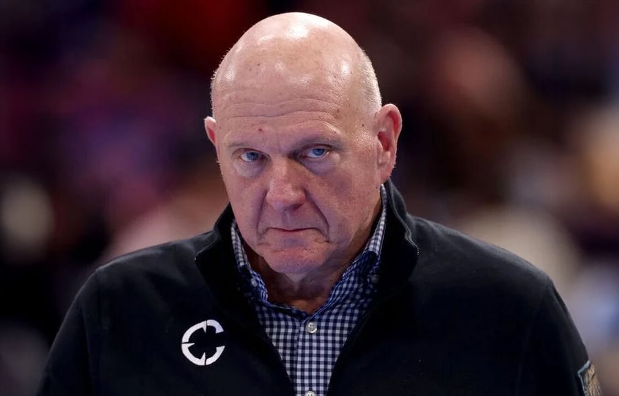 A bald man in a black jacket and checkered shirt looks forward with a serious expression. The background is blurred.