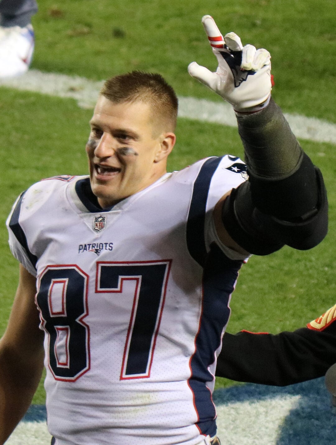 A football player wearing a New England Patriots jersey with the number 87 smiles and raises his gloved hand, making a gesture, on the field during a game.